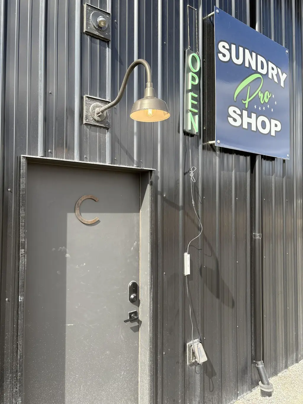 Sundry Pro Shop entrance in Columbia Falls, MT — blue Sundry Pro Shop sign and neon OPEN sign beside door C on the dark metal building