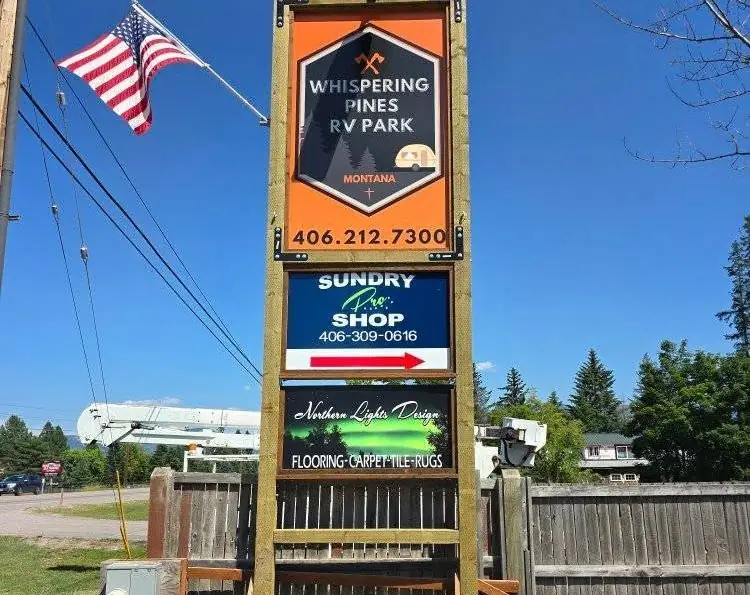 Sundry Pro Shop roadside sign on Highway 2 in Columbia Falls, Montana, below the Whispering Pines RV Park sign and above the Northern Lights Design sign